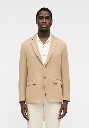 Homme noir portant un blazer beige sur une chemise blanche et un pantalon clair, debout devant un fond blanc uni.