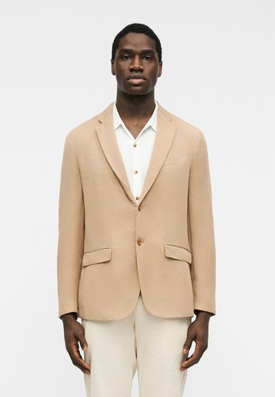 Homme noir portant un blazer beige sur une chemise blanche et un pantalon clair, debout devant un fond blanc uni.