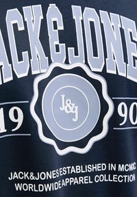 Camiseta azul marino con un gran logo en blanco y azul claro con diseño circular, texto "JACK&JONES" y gráficos del año de establecimiento.