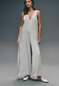 Ljusgrå ärmlös jumpsuit med djup V-ringning, flödande tyg och vida ben. Har en jämn textur och minimalistiska designinslag.