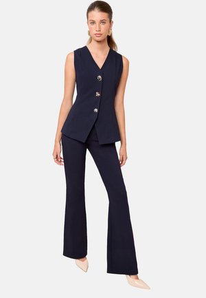 Modella che indossa un gilet blu navy con bottoni dorati e pantaloni a campana coordinati, in piedi con sicurezza su uno sfondo semplice.