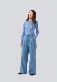 Blå mønstrete langermet skjorte sammen med høyhånds, vide lys blå jeans. Modellen står med den ene hånden i lommen.