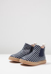 Bottes pour tout-petits bleu marine avec des pois blancs, semelles beige, fermeture éclair sur le côté et panneaux élastiques sur une surface blanche.