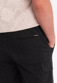 Pantalons cargo noirs avec une poche latérale, fabriqués en matériau durable. La taille est élastique pour le confort et comporte une petite étiquette de marque.