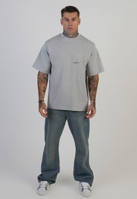 Pelēks kokvilnas t-krekls ar īsām piedurknēm un apaļu kakla izgriezumu, ar mazu melnu logotipu uz krūts. Nēsā ar brīviem ziliem džinsiem un baltām sporta apaviem.