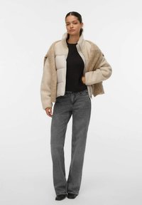Kvinde iført beige sherpa- og quiltet jakke over sort sweater med grå højhalset straight-leg jeans og sorte ankelstøvler, stående.