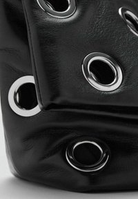 Bolso de cuero negro con ojales plateados, textura suave y un diseño plegado, que destaca detalles de hardware en contraste.