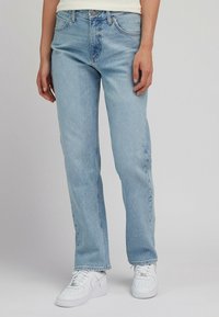 Jeans de denim azul claro con corte recto, que cuentan con cinco bolsillos, cremallera y un ligero desgaste para textura. Combinados con zapatillas blancas.