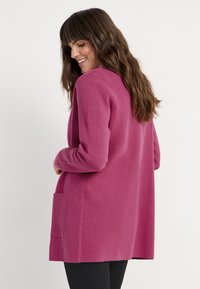 Långärmad cardigan i livlig rosa, med en texturerad stickning, öppen fram och en ficka på nedre vänstra sidan.