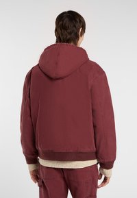 Person vist fra ryggen iført en bordeauxfarvet hættetrøje, matchende bukser og en beige strikket trøje synlig ved håndleddene og i kanten.