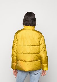 Gele pufferjas met een glanzende afwerking, voorzien van een hoge kraag en een horizontaal doorgestikt patroon. Achteraanzicht toont korte mouwen en een ontspannen pasvorm.