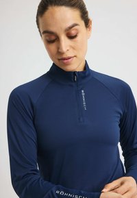 Frau in einem navyblauen Sportoberteil mit langen Ärmeln und Reißverschluss, auf dem "Röhnisch" auf dem Reißverschluss und dem Ärmel steht, schaut nach unten.