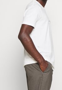 Camiseta de algodón blanca con mangas cortas y un ajuste relajado, combinada con pantalones de cuadros marrones. Detalle de marca sutil cerca del dobladillo.
