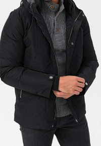 Sort pufjakke med hætte og lynlås. Har en tekstureret grå sweater under, knapdetaljer og tætsiddende manchetter.