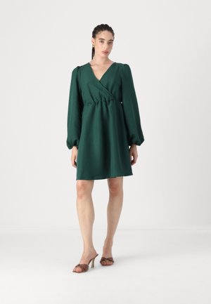 Green wrap dress made of textured fabric, featuring long puff sleeves, a cinched waist, and a mid-thigh hemline. Worn with brown heeled sandals.