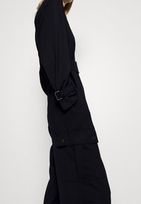 Vestido negro de manga larga con cinturón en la cintura, que cuenta con una tela lisa, grandes bolsillos con cierre de botón y una silueta ajustada.
