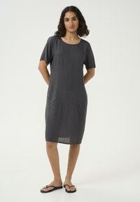 Robe en lin gris avec des manches courtes, un col rond et des poches latérales, présentant une coupe ample, tombant juste au-dessus des genoux. Associée à des tongs.