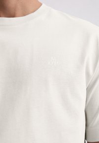 Hellgraues Baumwoll-T-Shirt mit Rundhalsausschnitt, dezent bestickt mit "M OP" auf der linken Brust, glatter Textur und einfachem Design.