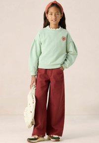 Sweatshirt vert menthe avec des manches bouffantes, col blanc à volants, patch en forme de cœur rouge. Pantalon bourgogne à jambes larges, tenant un sac crème avec des cœurs colorés.