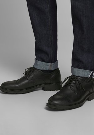 Des chaussures brogues noires en cuir avec des lacets portées avec un jean foncé à revers sur une surface grise.