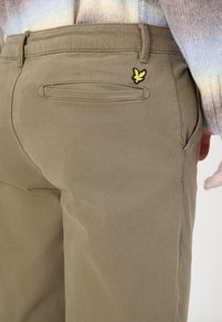 Khaki cotton trousers with a close-fit design, featuring a back pocket and a small yellow and black logo accent on the upper right.