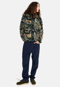 Giacca in pile con motivo camouflage verde e accenti arancioni e viola, dotata di zip completa e colletto alto. Abbinata a pantaloni blu navy e scarpe marroni.