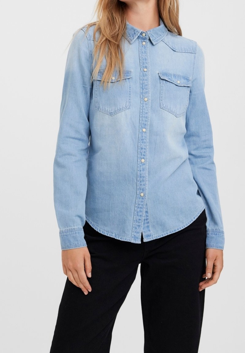 Camicia di jeans azzurro chiaro con maniche lunghe, colletto e due tasche sul petto. Orlo arrotondato e chiusura frontale con bottoni. Tessuto morbido con aree sbiadite.