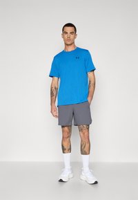 Blå kortermet skjorte med vertikal ribbet tekstur og svart logo, kombinert med grå shorts, båret med hvite joggesko og hvite sokker.