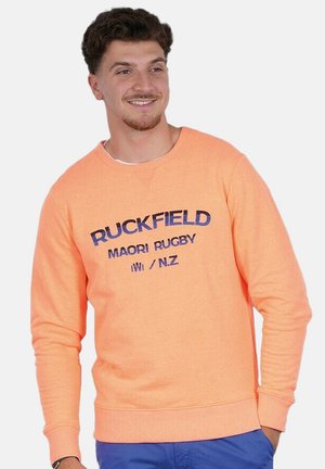 Homme souriant, portant un sweat-shirt orange avec le texte "RUCKFIELD MAORI RUGBY IWI/N.Z" et un pantalon bleu, sur un fond uni clair.