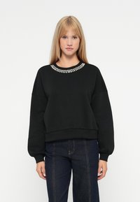 Sudadera negra con cuello redondo adornado, hombros caídos y puños acanalados. Presenta un ajuste corto y una textura suave.