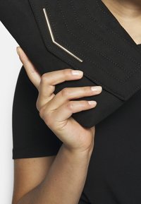 Černá semišová kabelka na rameno s diagonálním prošíváním a zlatým akcentem ve tvaru chevronu, držena jednou rukou na černém pozadí.