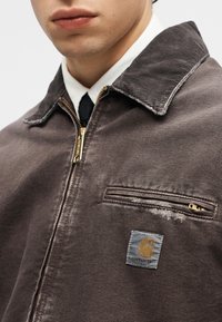 Άνδρας που φοράει καφέ ζακέτα Carhartt με φερμουάρ πάνω από λευκό πουκάμισο και μαύρη γραβάτα, δείχνοντας τον γιακά της ζακέτας και την τσέπη στο στήθος με το λογότυπο.