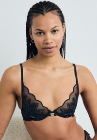 Μαύρο σουτιέν δαντέλας με ανθισμένα μοτίβα, οδοντωτές άκρες και ρυθμιζόμενους ιμάντες. Περιλαμβάνει μια μικρή ασημένια λεπτομέρεια στο κέντρο.