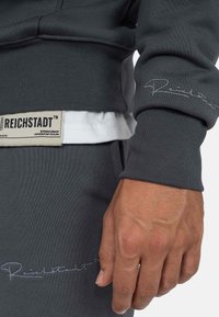 Felpa grigio scuro con polsini a coste, tessuto testurizzato e marchio ricamato lungo le maniche. Etichetta con la scritta "REICHSTADT."