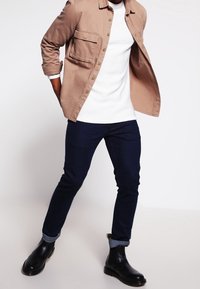 Camicia beige con bottoni e taschino sul petto, maglia bianca a coste sotto, jeans in denim scuri, maniche arrotolate e stivali Chelsea neri.
