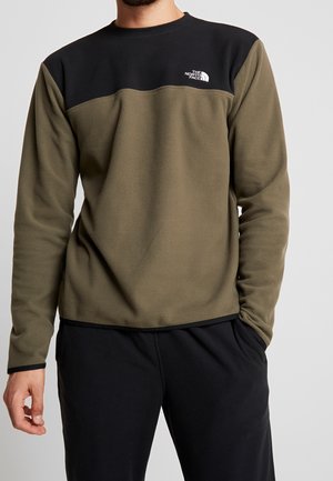 Homme portant un pull polaire olive vert et noir avec le logo The North Face sur la poitrine, associé à un pantalon noir, les mains détendues le long du corps.