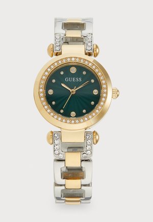 Une montre Guess aux tons dorés et argentés présente un cadran rond vert, des accents en cristal et un bracelet en métal mixte. Des embellissements semblables à des diamants ornent la lunette.