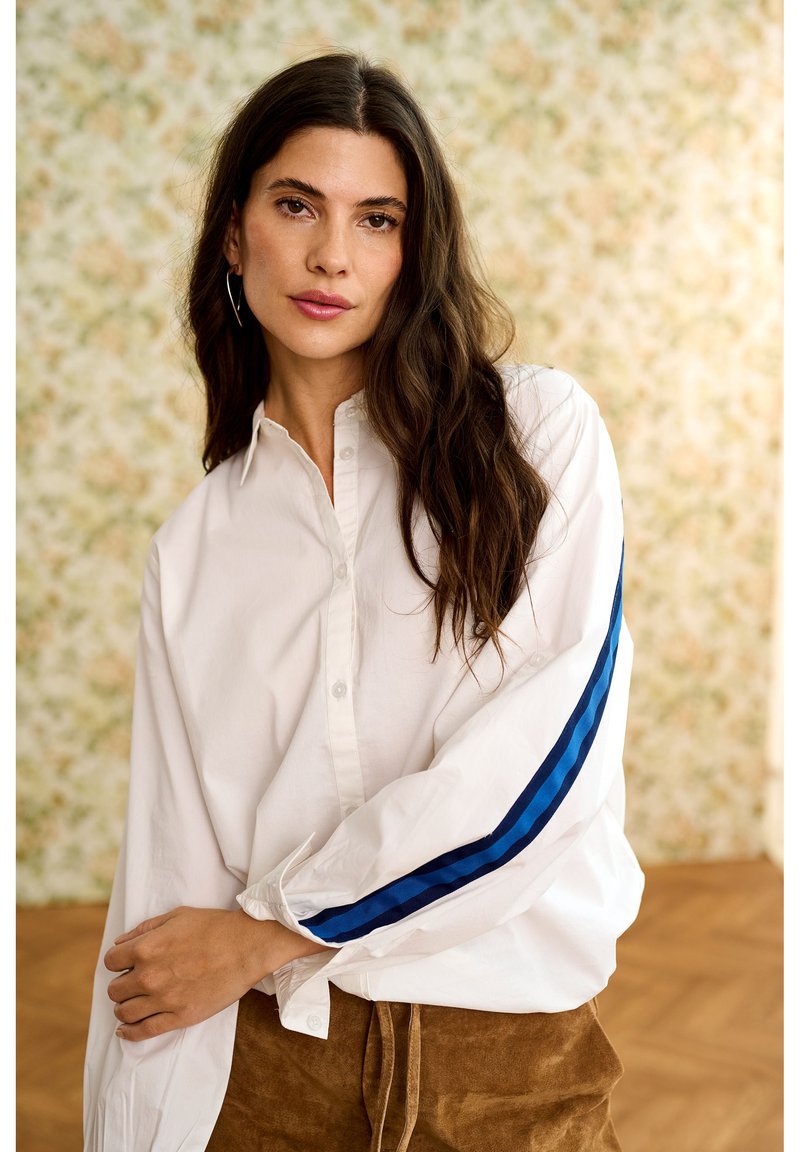 Femme aux longs cheveux foncés portant une chemise blanche boutonnée avec des rayures bleues sur les manches et un pantalon marron, debout à l'intérieur.