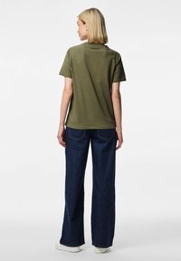 Lühikeste blondide juustega naine, kes kannab oliivrohelise värvi t-särki, tumedaid laiapõlvalisi teksaseid ja valgeid jalanõusid, seisab seljaga valge tausta ees.