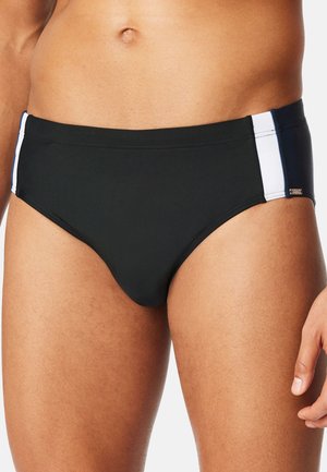 Nahaufnahme eines Mannes, der schwarze Badehosen mit weißen und marineblauen Seitenteilen trägt, wobei sein unterer Oberkörper und seine oberen Oberschenkel zu sehen sind.