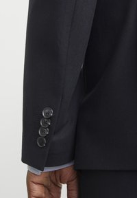 Ärmels eines Marine-Anzugsjackets mit vier schwarzen Knöpfen und subtilen Stickdetails, die einen blau-weiß gestreiften Bund darunter enthüllen.