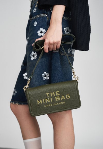 Cilvēks tur olīvzaļo ādas mini somu ar zelta burtiem "THE MINI BAG MARC JACOBS" blakus džinsa svārkiem ar sudraba ziedu aplikācijām.