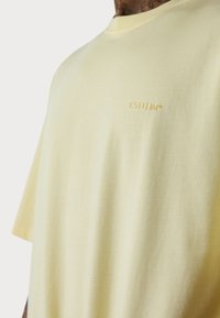 Kurze Ärmel, Rundhals-T-Shirt aus hellem gelbem Baumwollstoff, mit einem dezenten, bestickten Logo "ESTEEM" in passendem Faden auf der Vorderseite.