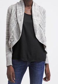 Ljust grå cardigan med öppen front, intrikata stickade mönster och fransdetaljer. Bärs över en svart topp och blå jeans. Mjuk textur.