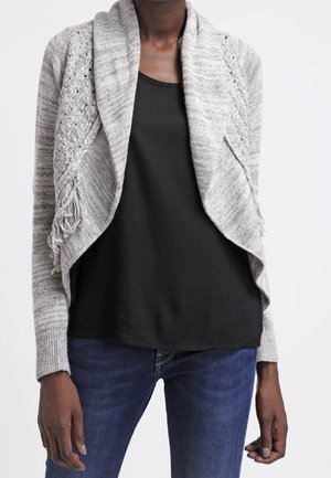 Cardigan gris clair avec une ouverture sur le devant, des motifs de tricot complexes et des accents en frange. Porté sur un haut noir et un jean bleu. Texture douce.