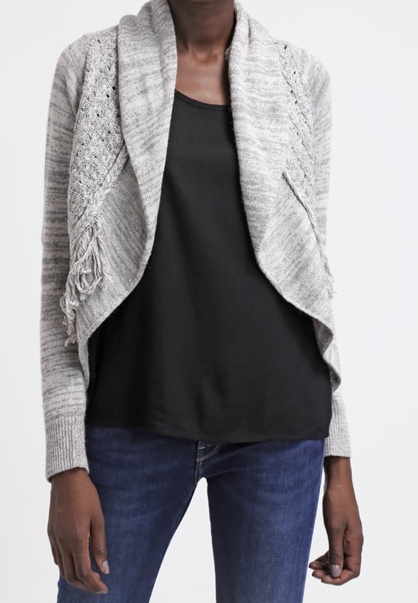 Ljust grå cardigan med öppen front, intrikata stickade mönster och fransdetaljer. Bärs över en svart topp och blå jeans. Mjuk textur.