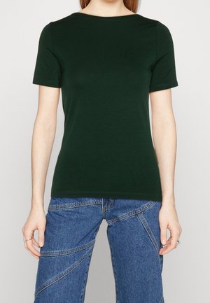 Kobieta w ciemnozielonym dopasowanym t-shircie z krótkim rękawem i niebieskich dżinsach z ukośnymi przeszyciami, stoi na jednolitym tle.
