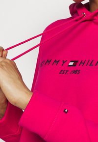 Jasnoróżowa bluza z kapturem z sznurkami, z haftowanym napisem "TOMMY HILFIGER" i logo. Mankiety ozdobione małą metką z logo. Miękka, fakturowana tkanina.