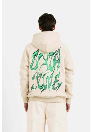 Beige hooded sweatshirt featuring a green graphic design on the back, with a fluid, wavy font pattern. Soft fabric with a relaxed fit.