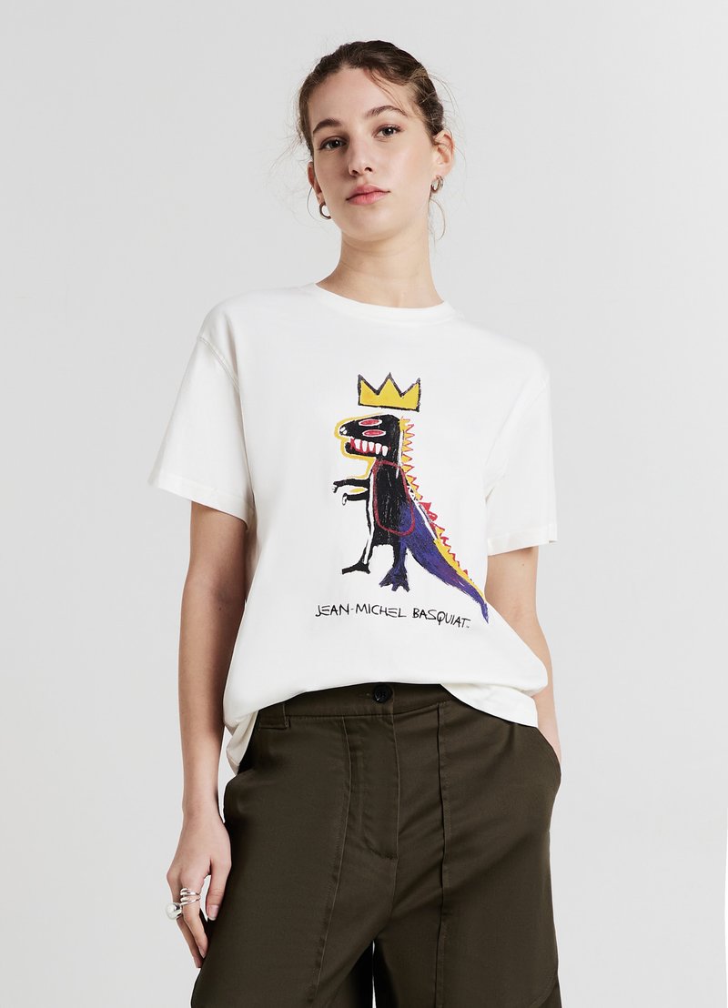 T-shirt en coton blanc avec un graphique coloré d'un dinosaure portant une couronne et le texte "JEAN-MICHEL BASQUIAT." Manches courtes, coupe décontractée.